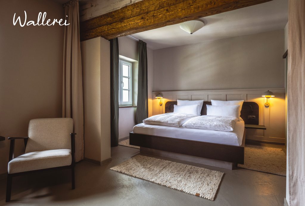Schlafzimmer im Hotel Wallerei mit Doppelbett, weißen Kissen, Holzbalken an der Decke und einem Sessel neben dem Fenster
