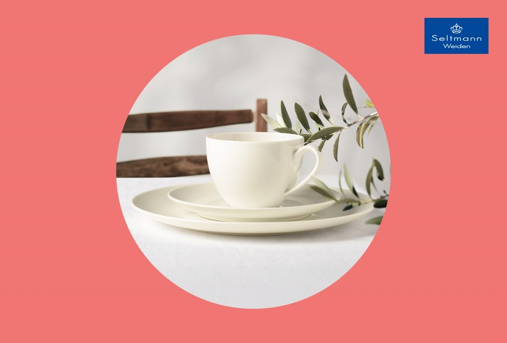 Weiße Porzellantasse mit Untertasse und Teller auf weißem Tisch, dahinter Holzstuhl und Olivenzweig, Seltmann Weiden Logo oben rechts