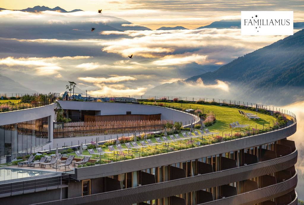 Hotelansicht des Hotels Familiamus mit Dachgarten unter bewölktem Himmel und Bergpanorama